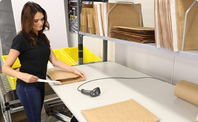 Une employée ferme une enveloppe matelassée en papier kraft dans un environnement d'expédition. Autour d'elle, des étagères qui contiennent plusieurs enveloppes prêtes à l'emploi.