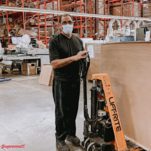 Un employé de SupremeX se tient dans un entrepôt, portant un masque et utilisant un transpalette manuel à côté d'une pile de matériaux. L'espace de travail comprend des étagères bien organisées et de l'équipement industriel en arrière-plan.