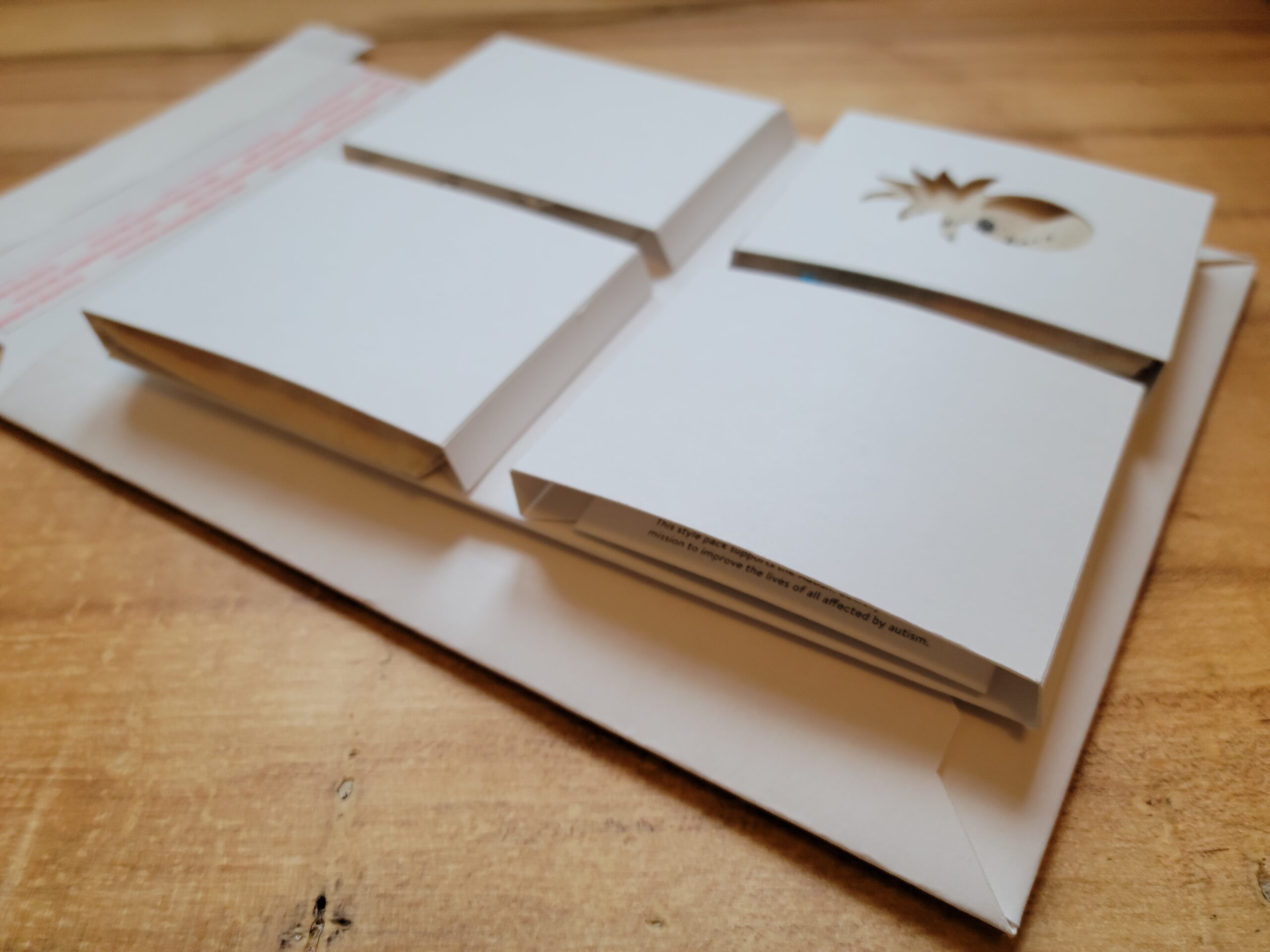 Une vue latérale d’un emballage personnalisé composé de plusieurs compartiments en carton blanc. L’un des compartiments présente une découpe en forme d’ananas, laissant entrevoir un design imprimé à l’intérieur. L’emballage est posé sur une enveloppe en carton avec une bande adhésive rouge, suggérant une solution d’expédition. La conception soignée et minimaliste met en valeur la hauteur et la structure des compartiments, indiquant une approche fonctionnelle et esthétique pour la présentation des produits.